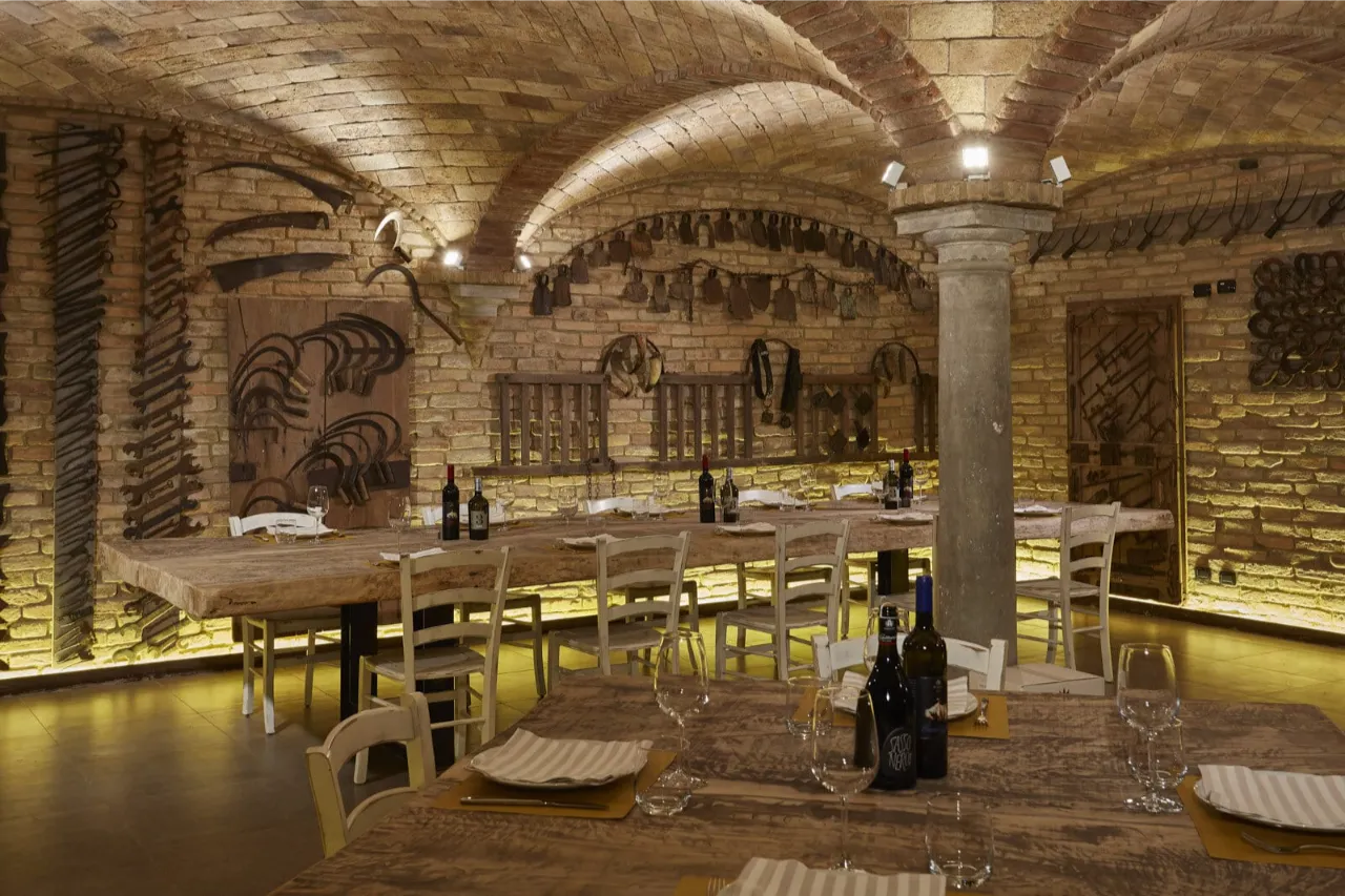 High-end rustic Italian restaurant interior in Fidenza featuring bespoke antique brick sail vaults (volte a vela). The warm, textured flat-laid bricks (pianelle) harmonize beautifully with stone columns, long rustic wooden dining tables, and a unique wall display of antique agricultural tools, creating a deeply authentic and elegant dining atmosphere.