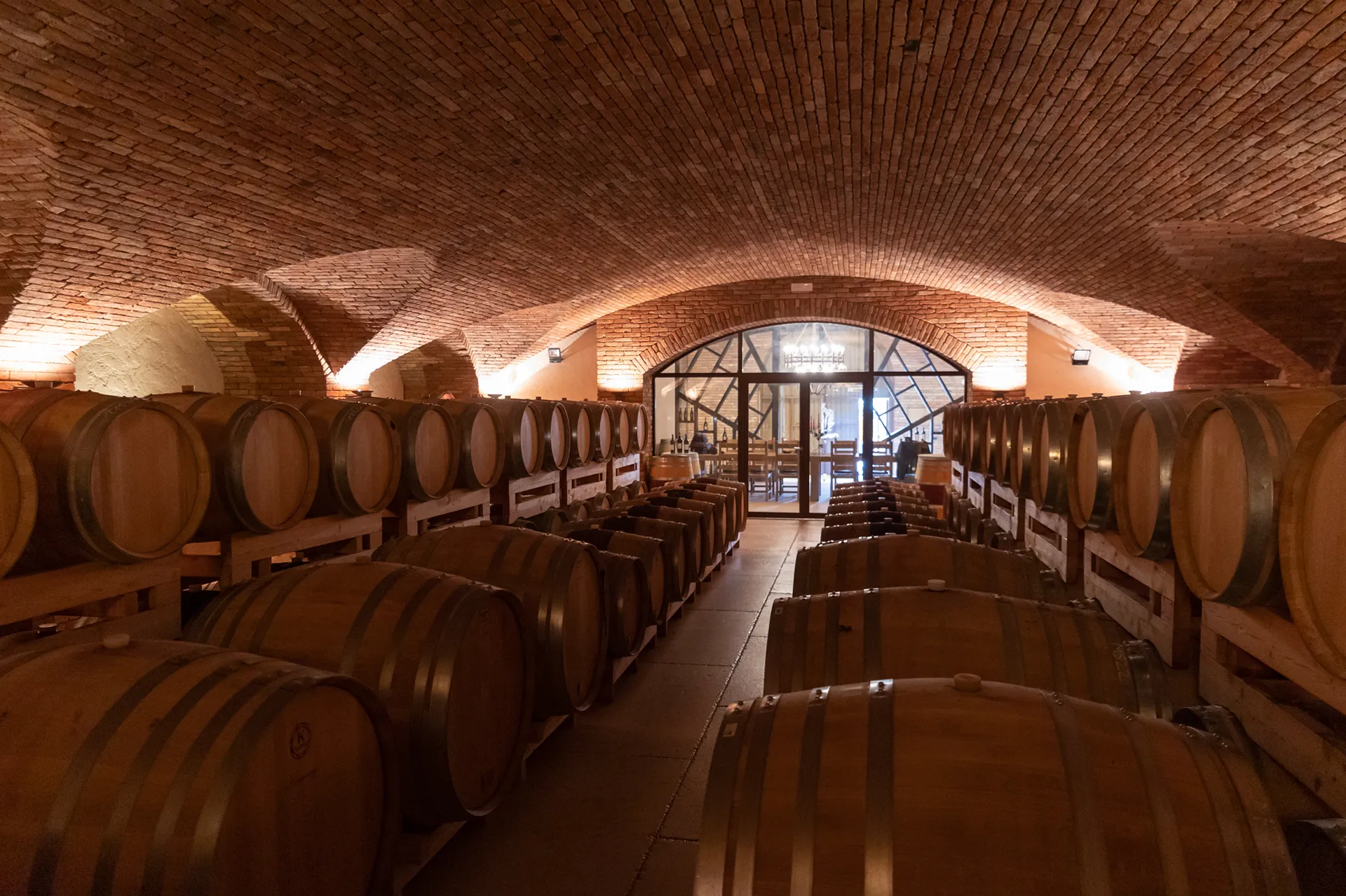 Expansive commercial wine cellar featuring masterful Italian masonry. A continuous brick barrel vault with lateral lunettes (unghie) creates a profound architectural rhythm, seamlessly connecting the barrel aging area to the tasting room through an elegant glass partition at Giusti Wine.
