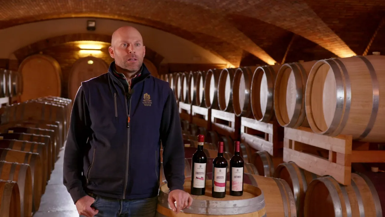 Video testimonial cover showing the expansive commercial wine cellar at Giusti Wine. Features a masterful continuous antique brick barrel vault with lateral lunettes (unghie), creating an elegant and profound architectural rhythm for the barrel aging room.