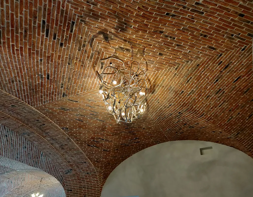 Designer chandelier against an antique brick vaulted ceiling, warm light grazing the texture — by Pietrantiche